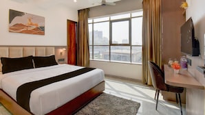 Ein Hotelzimmer mit einem großen Bett, einem Schreibtisch mit Stuhl, einem Flachbildfernseher, einem Fenster mit Vorhängen und einem Bild an der Wand.