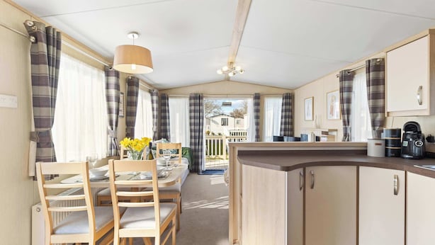A compact kitchen with wooden cabinets, a dining area with chairs, a table, and a view of a house through the window. - Charming Beachside Chalet close to clubhouse (Pevensey Bay)