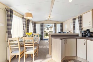 Een kleine keuken met houten kastjes, een eethoek met stoelen en een tafel, en uitzicht op een huis door het raam.