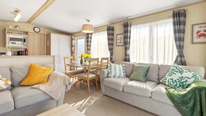 A cozy living room with a sofa, a dining table with chairs, a kitchen area with a microwave, and a window with curtains. - Charming Beachside Chalet close to clubhouse (Pevensey Bay)
