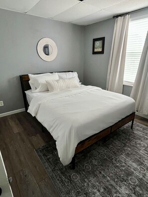 A neatly made bed in a room with a gray wall, a round mirror, and a framed picture.
