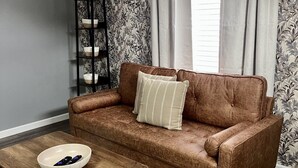 A brown leather sofa with a white pillow, a wooden coffee table with a bowl of remotes, a patterned wallpaper, and a window with curtains. - Newly Furnished St. Charles Getaway- B (Saint Charles)