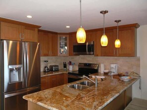 A modern kitchen with stainless steel appliances, granite countertops, wooden cabinets, and pendant lighting. - NEW! Peaceful Ocean-View 2BR Loft Escape - Unit C38 (Lahaina)