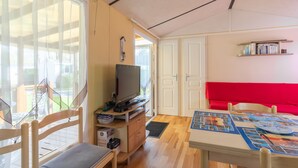 A room with a television on a wooden stand, a red sofa, a table with a board game, and a bookshelf. - Mobile Home Near Royan with Shared Pool, Private Terrace, and Wi-Fi (Meschers-sur-Gironde)