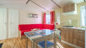 A compact room with a kitchenette, a dining table with a placemat, a red sofa, and a small bookshelf. - Mobile Home Near Royan with Shared Pool, Private Terrace, and Wi-Fi (Meschers-sur-Gironde)