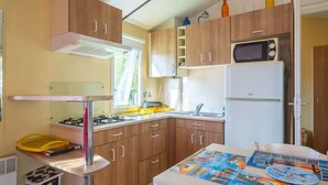 A compact kitchen with wooden cabinets, a microwave, a refrigerator, a gas stove, and a sink. - Mobile Home Near Royan with Shared Pool, Private Terrace, and Wi-Fi (Meschers-sur-Gironde)
