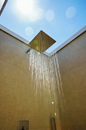 Une douche où l’eau s’écoule d’un pommeau carré fixé au plafond, le tout adossé à un mur, avec un ciel bleu dégagé au-dessus.