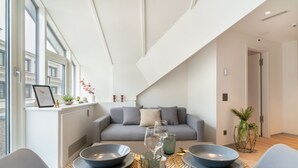 Uma sala de estar moderna com um sofá cinza, uma mesa de jantar posta com pratos, copos e talheres, e vista para o exterior através de grandes janelas.