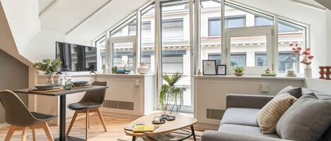 A modern loft apartment with a dining area, a sofa, and a large window offering a city view.