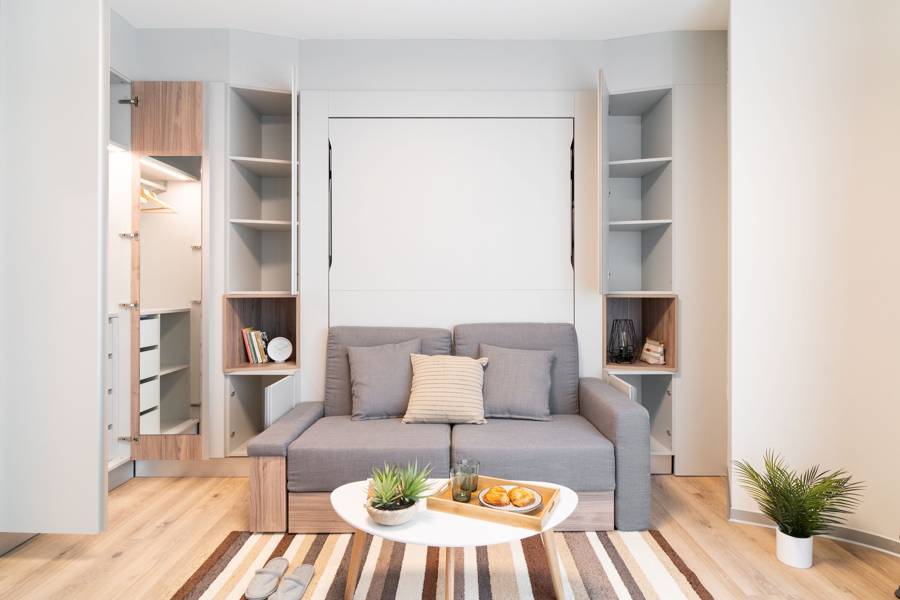 Uma sala de estar moderna com um sofá cinzento, uma mesa de centro com objetos decorativos, um tapete listrado e um chão de madeira.