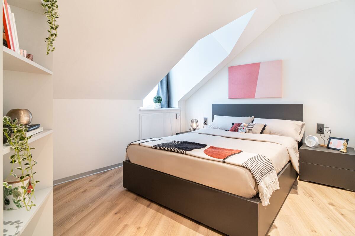 A bedroom with a bed, bedside tables, a nightstand, a wall-mounted shelf, and a hanging plant.