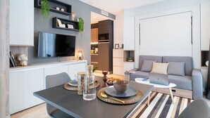 Uma sala de estar moderna com um sofá cinzento, uma mesa de jantar com jogos americanos, uma TV fixada na parede e uma área de cozinha ao fundo.
