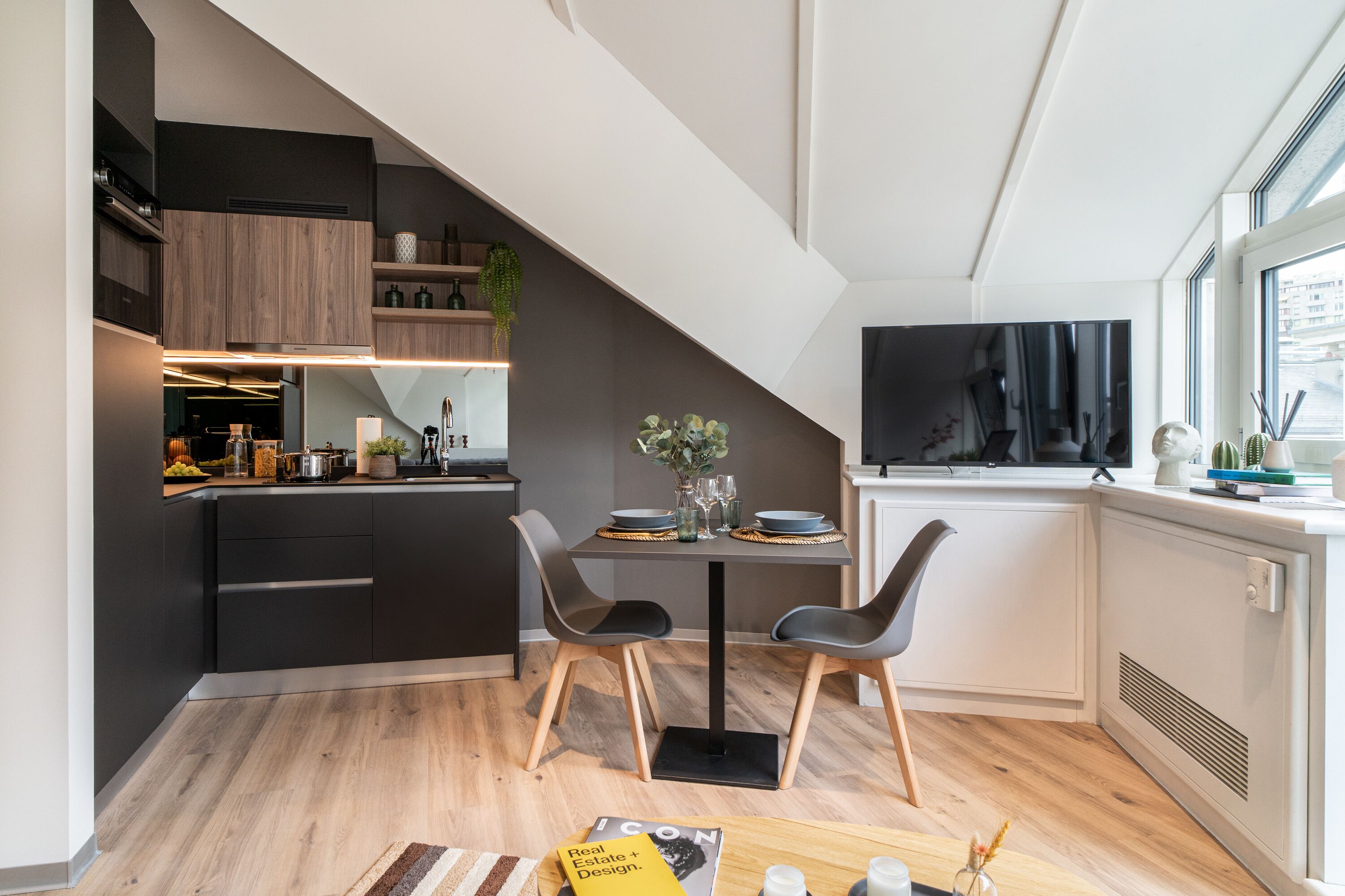 A modern kitchen with dark cabinets, a dining area with a table set for two, and a large window with a city view.