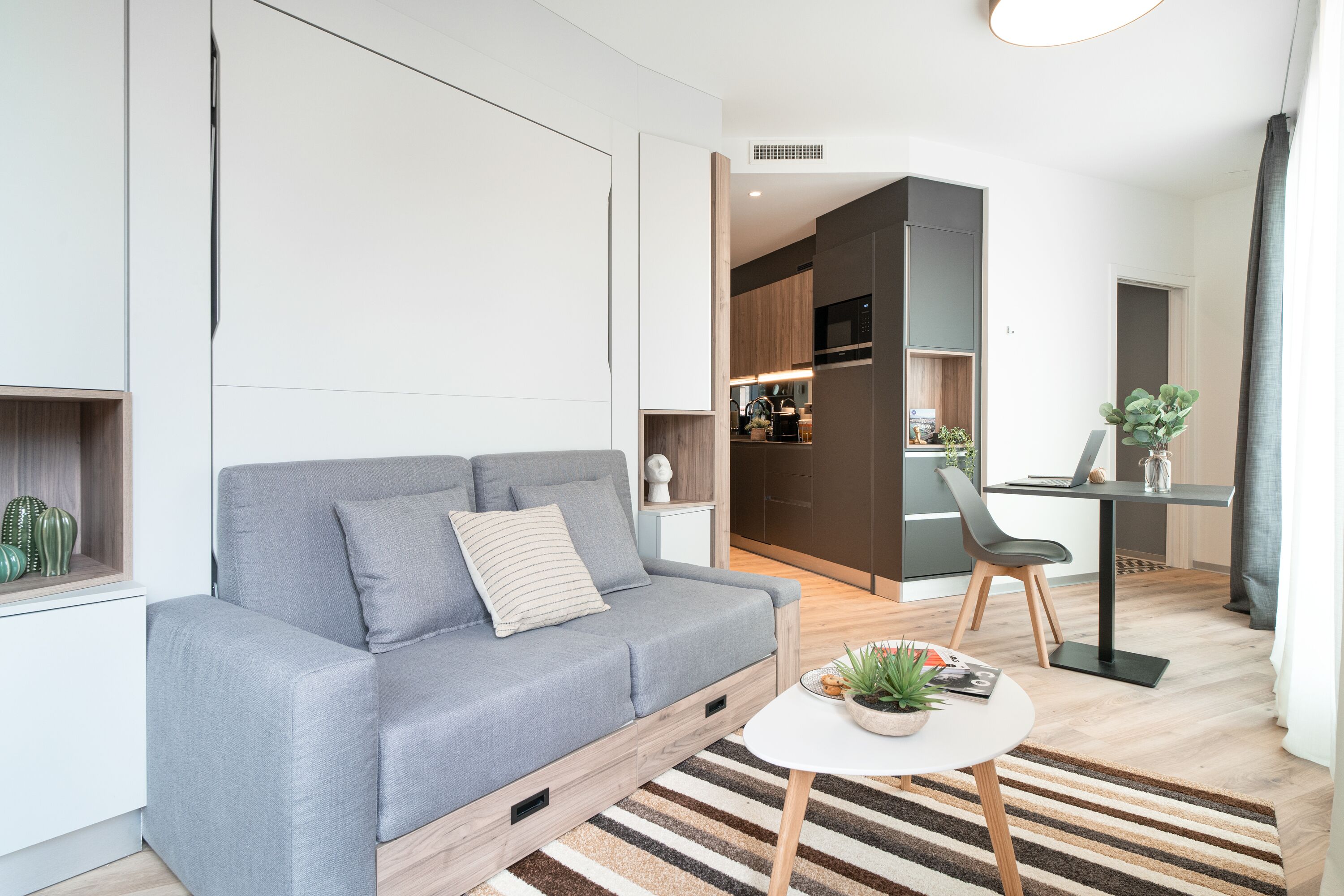 A modern living room with a grey sofa, a small round table with a plant, and a kitchen area in the background.