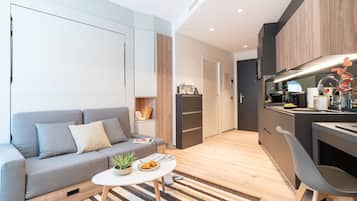 A modern living room with a grey sofa, a small round table with a plant and a plate of food, a striped rug, and a kitchen area with wooden cabinets and a sink.