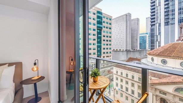 Un balcon donnant sur les bâtiments de la ville, une petite table avec une lampe, une chaise et une plante en pot.