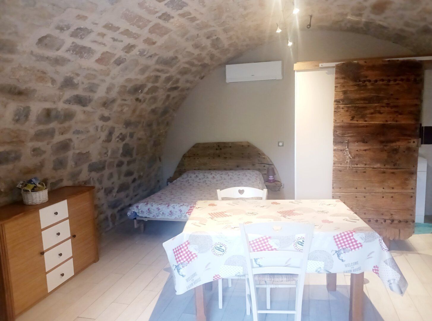 Een rustieke kamer met een stenen muur, een bed, een houten dressoir en een tafel met een geruit tafelkleed.