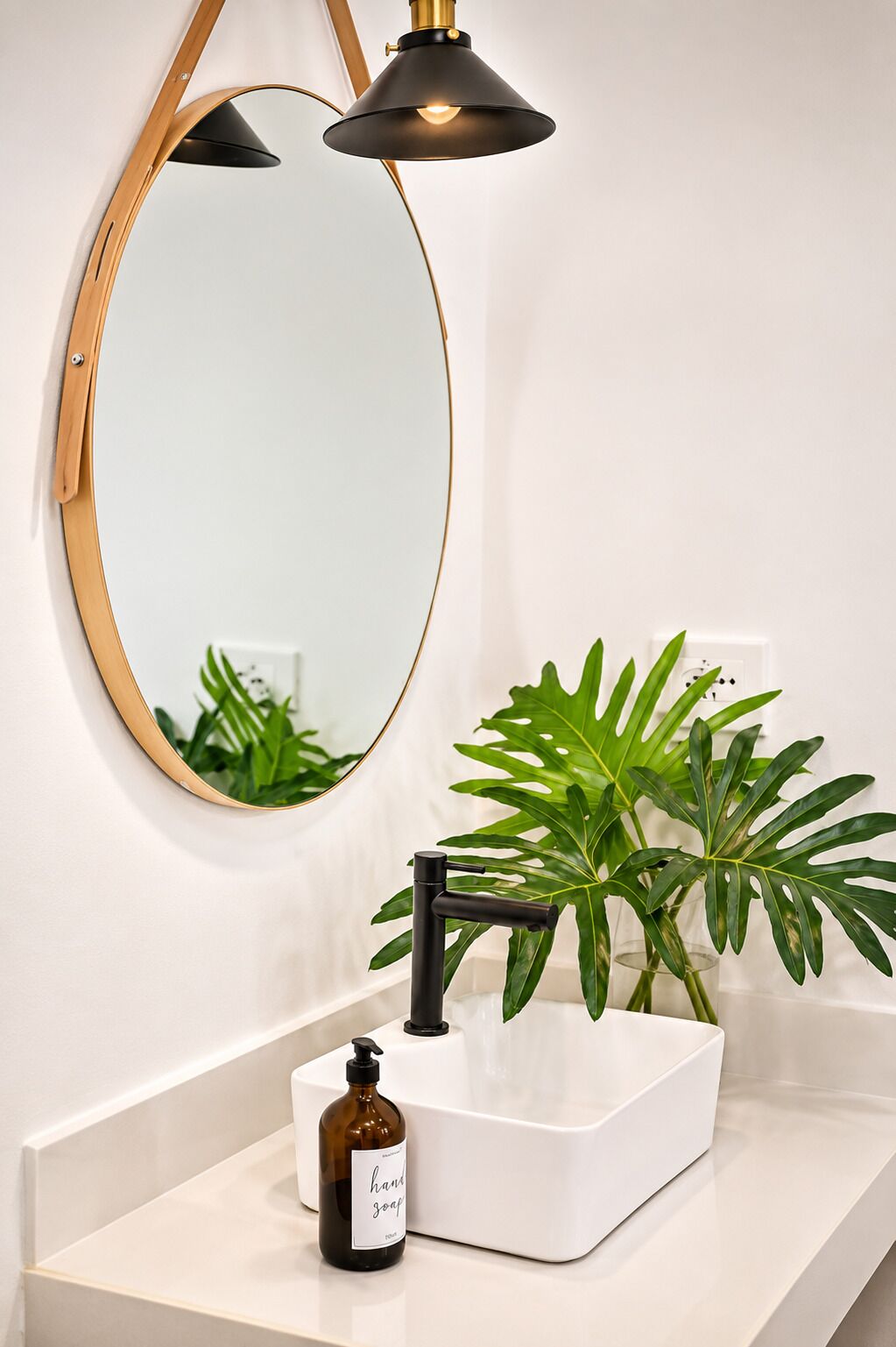Een badkamer met een witte wastafel, een zwarte kraan, een ronde spiegel met gouden lijst en een plant in pot.