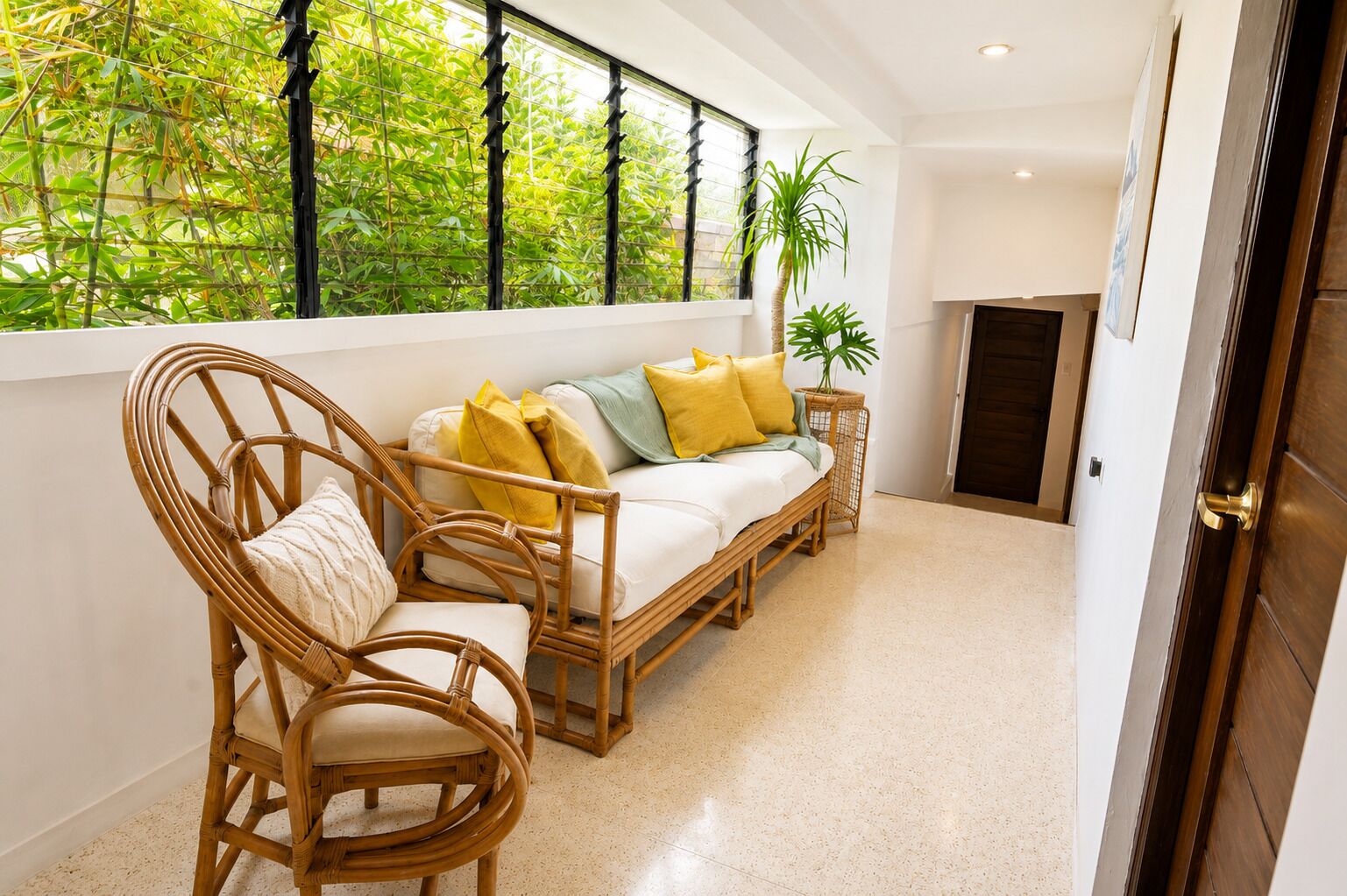 A room with a sofa, two wicker chairs, a small side table, a potted plant, and a view of greenery through a window.