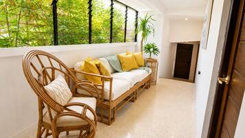 A room with a sofa, two wicker chairs, a small side table, a potted plant, and a view of greenery through a window.