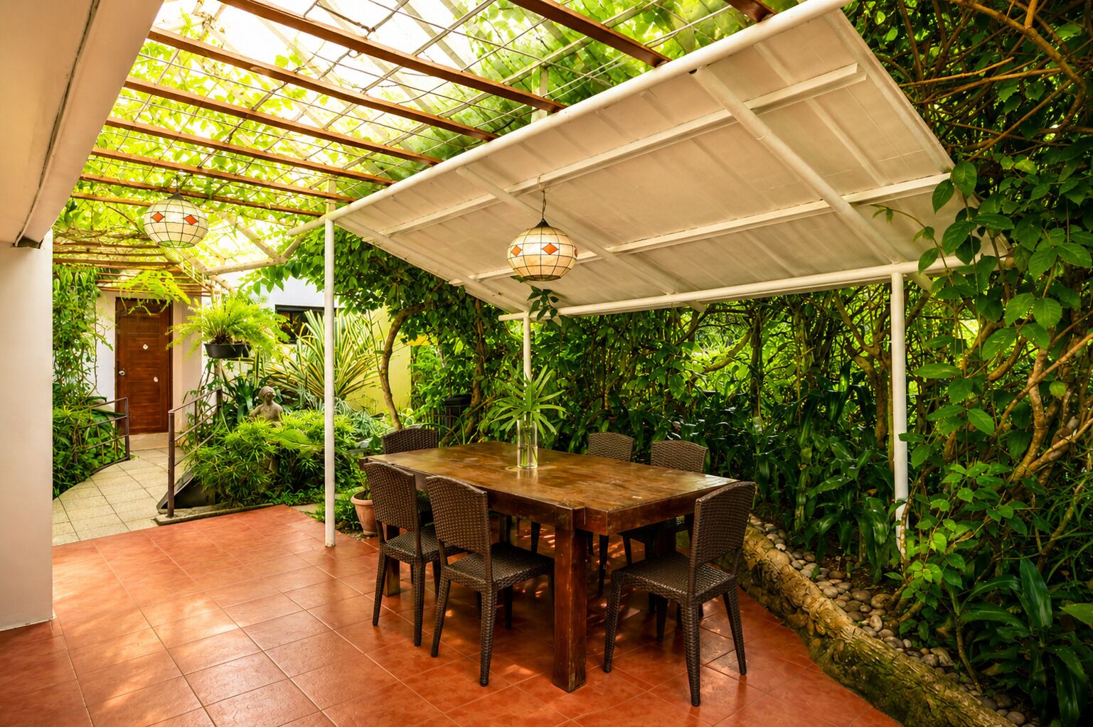 Un patio al aire libre con una mesa de madera y sillas de mimbre, rodeado de exuberante vegetación y una pérgola blanca.