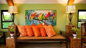 Een kamer met een houten bank, oranje kussens, een kleurrijk papegaaien schilderij en twee lampen.