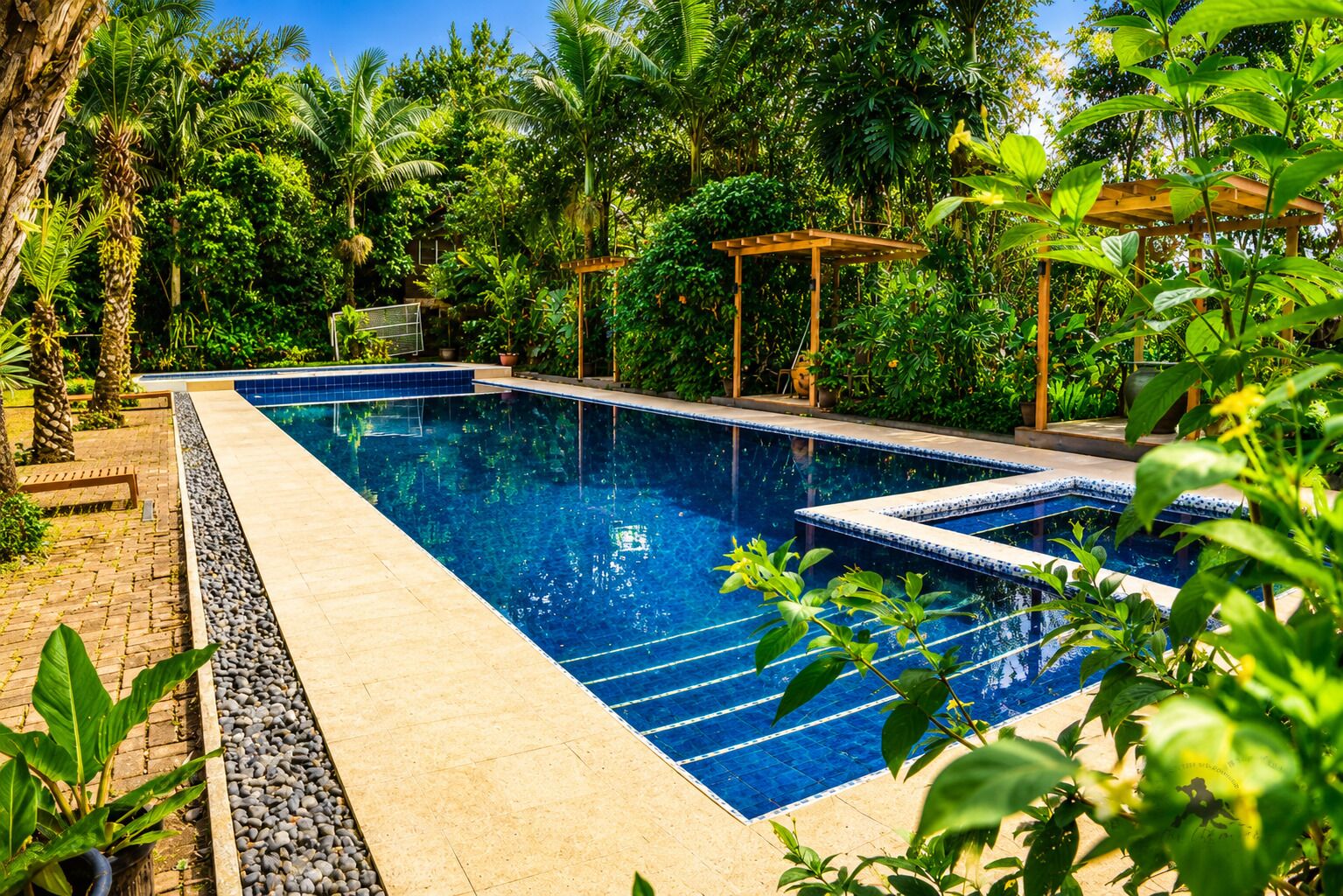 A rectangular swimming pool with clear blue water, surrounded by lush greenery and a wooden pergola.