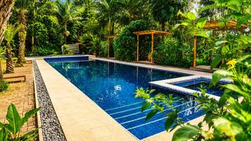 A rectangular swimming pool with clear blue water, surrounded by lush greenery and a wooden pergola.