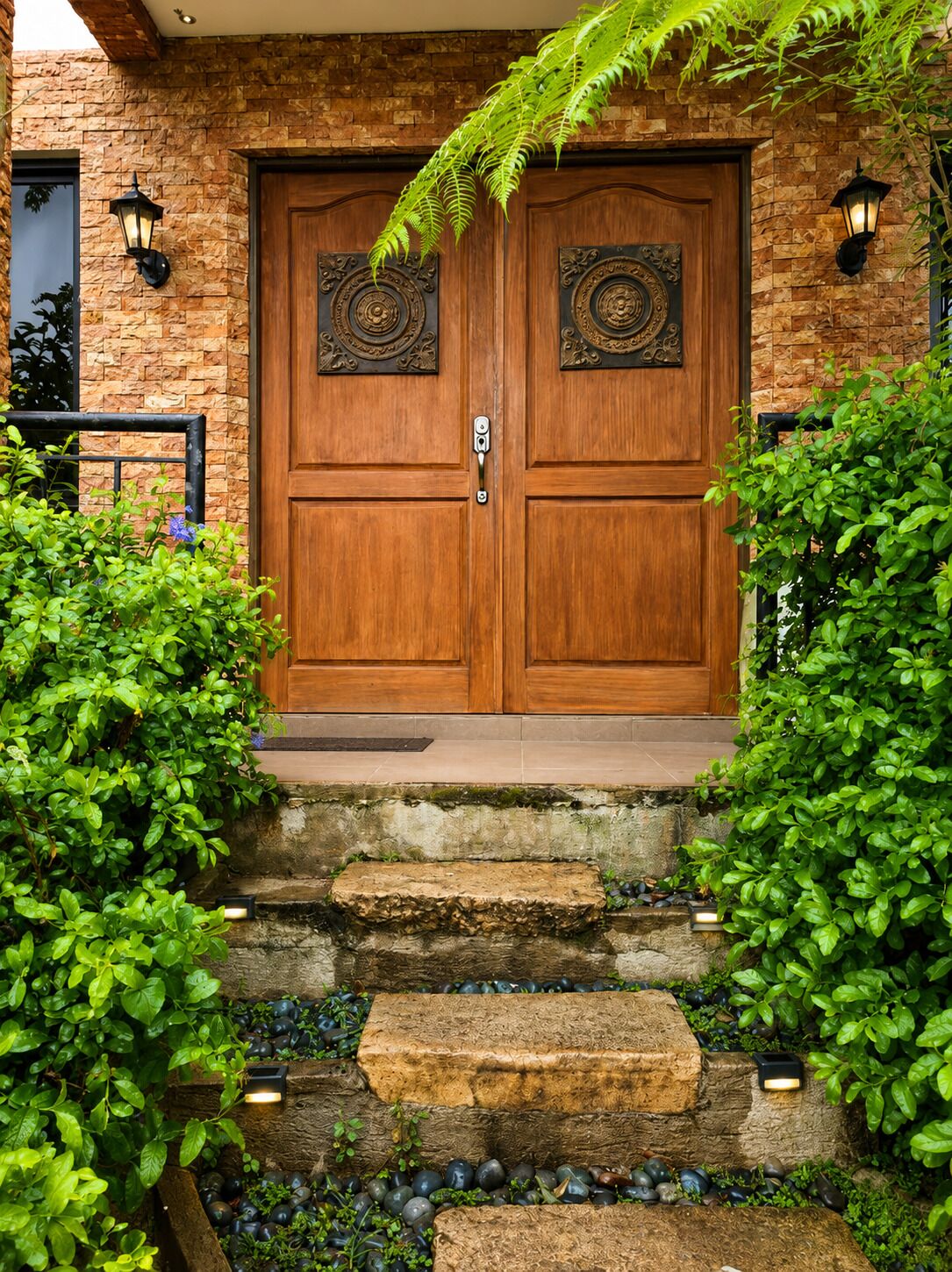 A wooden double door with decorative metalwork, flanked by lush green plants and a stone staircase leading up to the entrance.