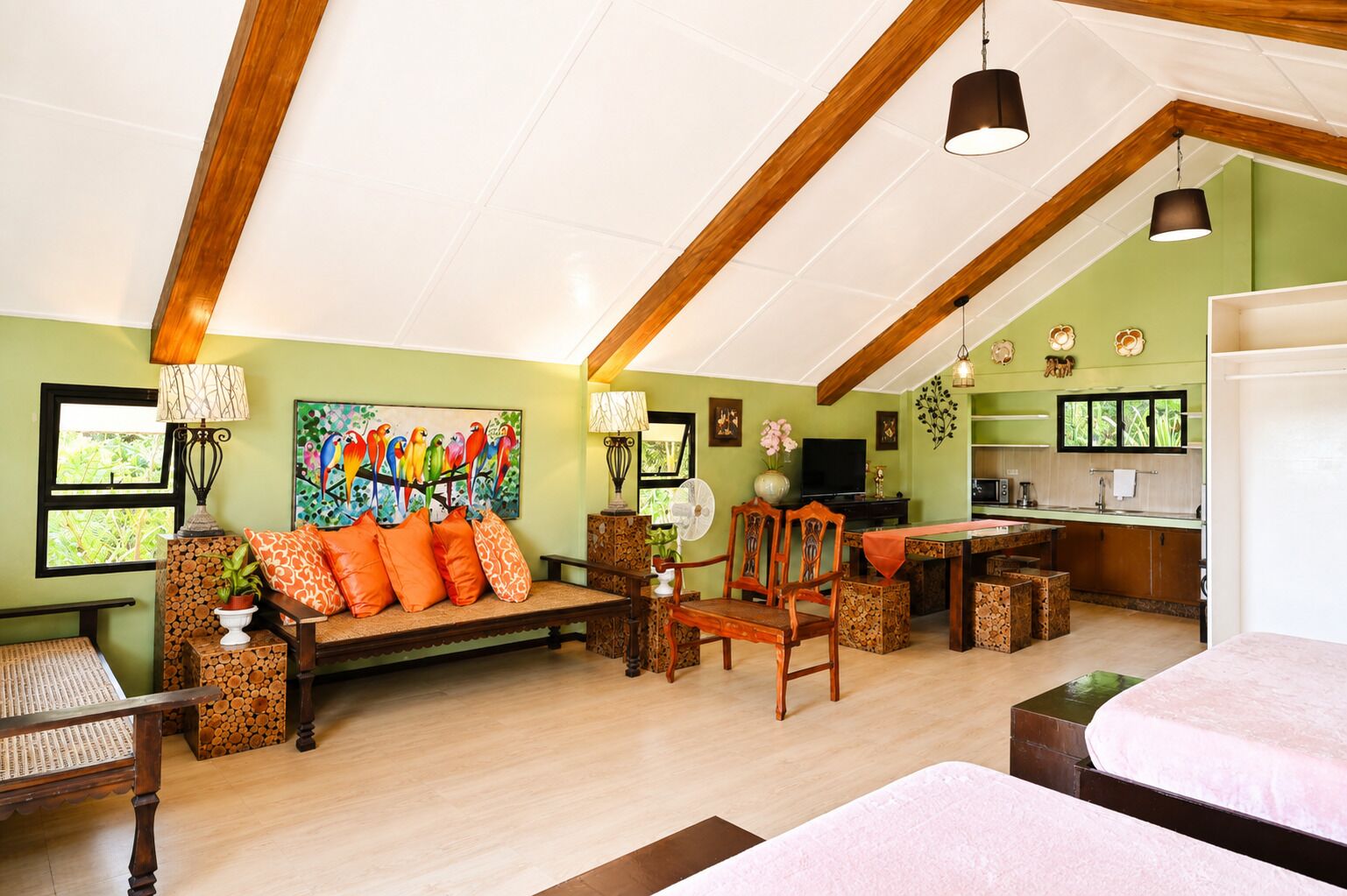 A spacious living area with a wooden ceiling, a sofa with orange cushions, a dining table, and a kitchen area with a TV and cabinets.