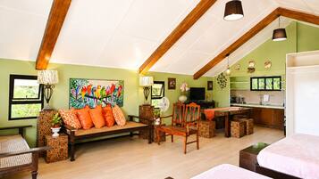 A spacious living area with a wooden ceiling, a sofa with orange cushions, a dining table, and a kitchen area with a TV and cabinets.