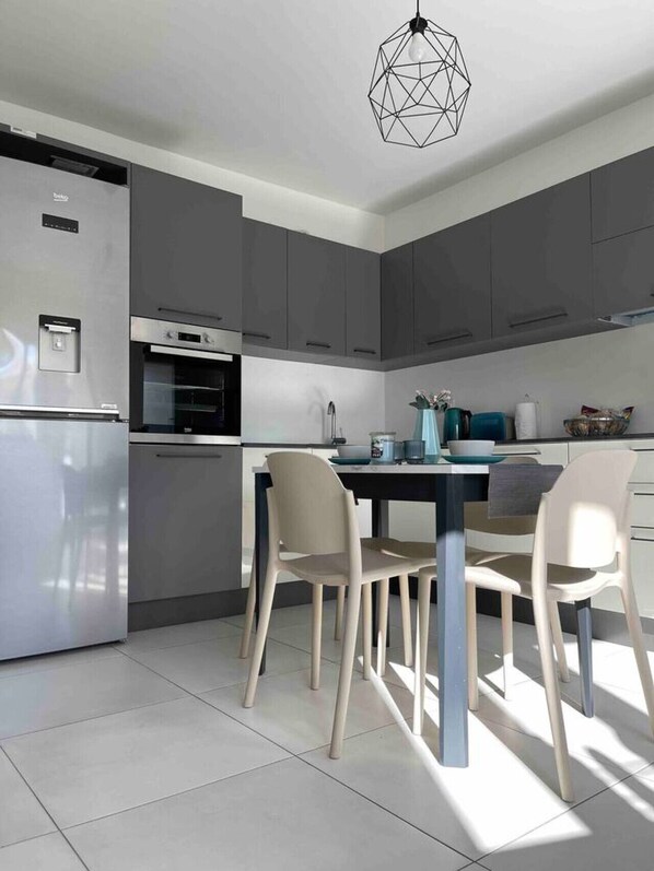 A modern kitchen with grey cabinets, a stainless steel refrigerator, a built-in oven, and a dining area with a table and chairs. - Zen Apt in the Heart of Malta (Ta` Xbiex)