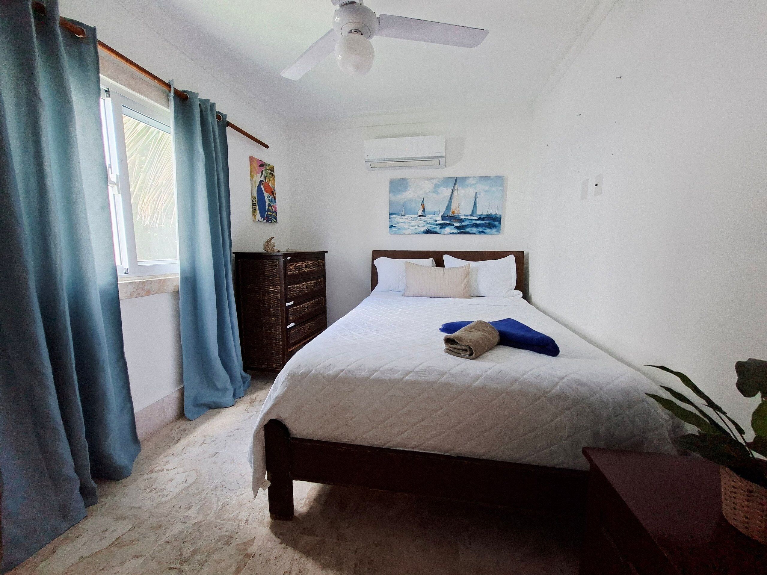 A bedroom with a bed, a ceiling fan, a window with curtains, a dresser, and a painting on the wall.