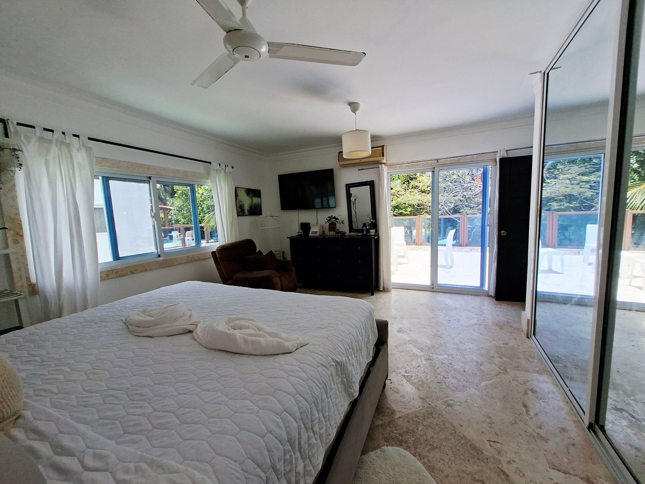 Ein Schlafzimmer mit Bett, Deckenventilator, Kommode, Fernseher und einer Schiebetür zu einem Außenbereich.