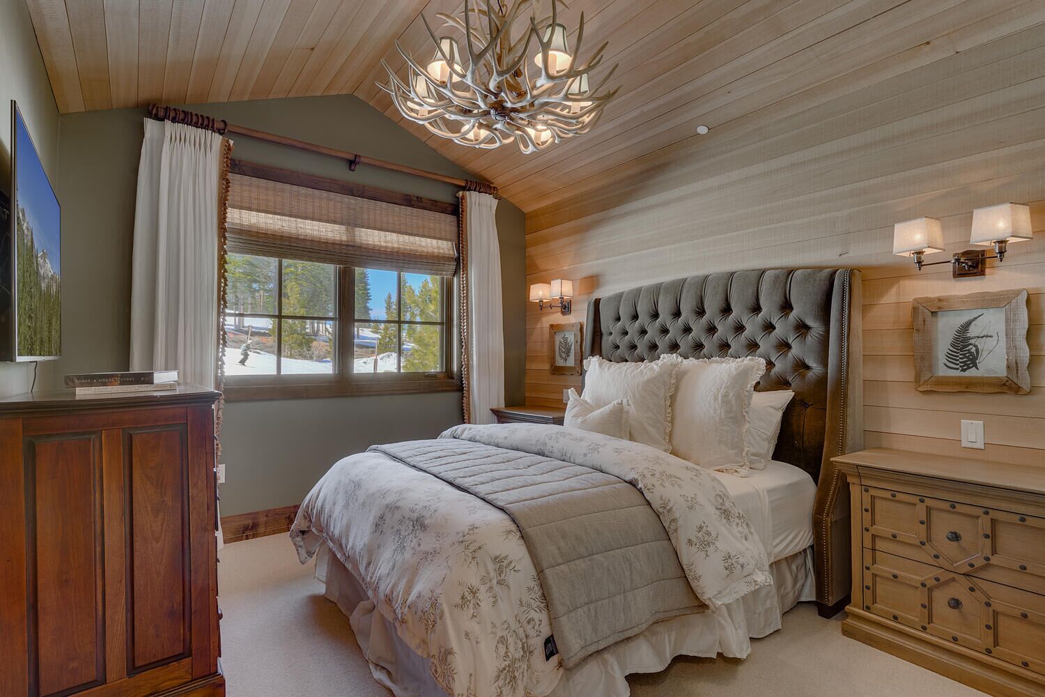 A bedroom with a tufted headboard, a large window with curtains, a wooden ceiling, and a chandelier.