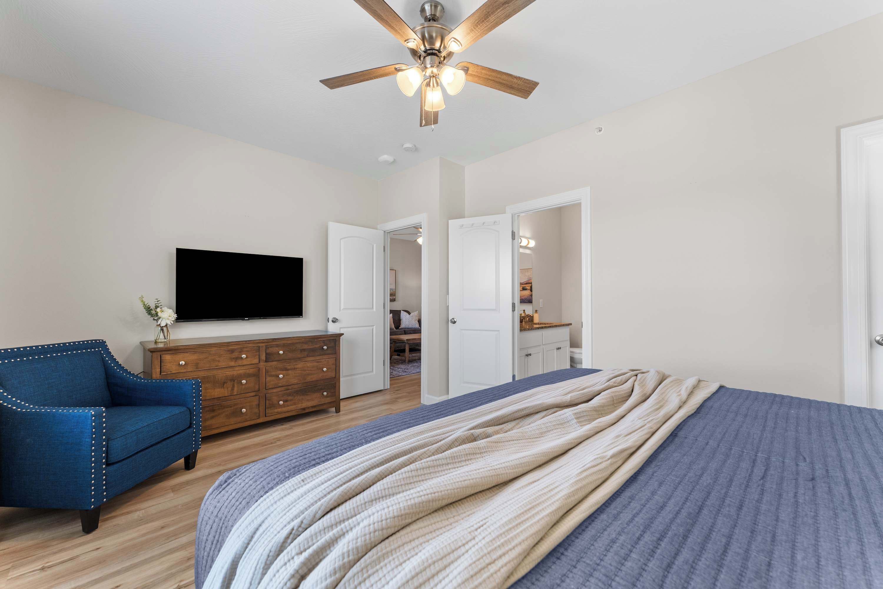A bedroom with a bed, a television, a ceiling fan, a dresser, and a blue armchair.