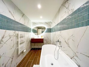 Une salle de bain avec un mur en marbre, un plancher en bois, une baignoire blanche et un miroir doté d’un cadre circulaire.