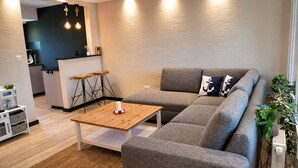 Ein modernes Wohnzimmer mit einer grauen Couch, einem Holztisch und einem kleinen Küchenbereich im Hintergrund.