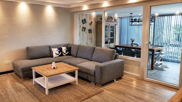 Ein modernes Wohnzimmer mit einer Eckcouch, einem Holztisch und einer Schiebeglastür, die zu einem Außenbereich führt.