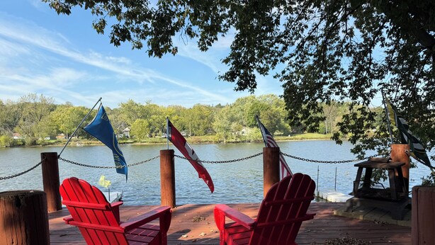 A wooden dock with two red Adirondack chairs facing a lake, surrounded by chains and flags.