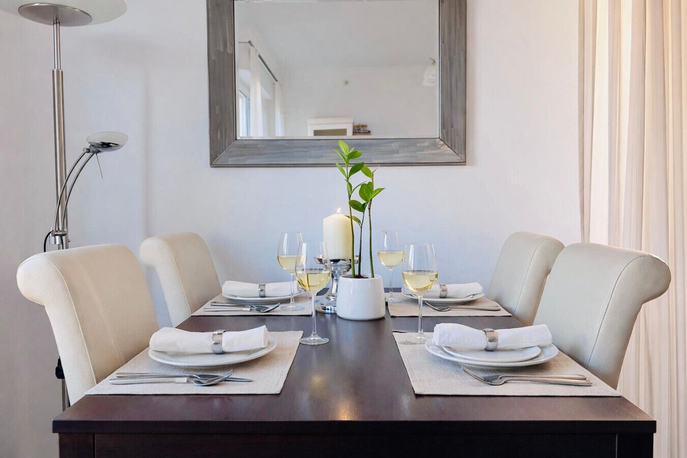 A dining table set for four with white plates, silverware, and glasses, flanked by two chairs, a candle centerpiece, and a mirror above.