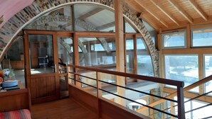 A spacious loft with a high ceiling, wooden beams, and large windows offering views of the outdoors.
