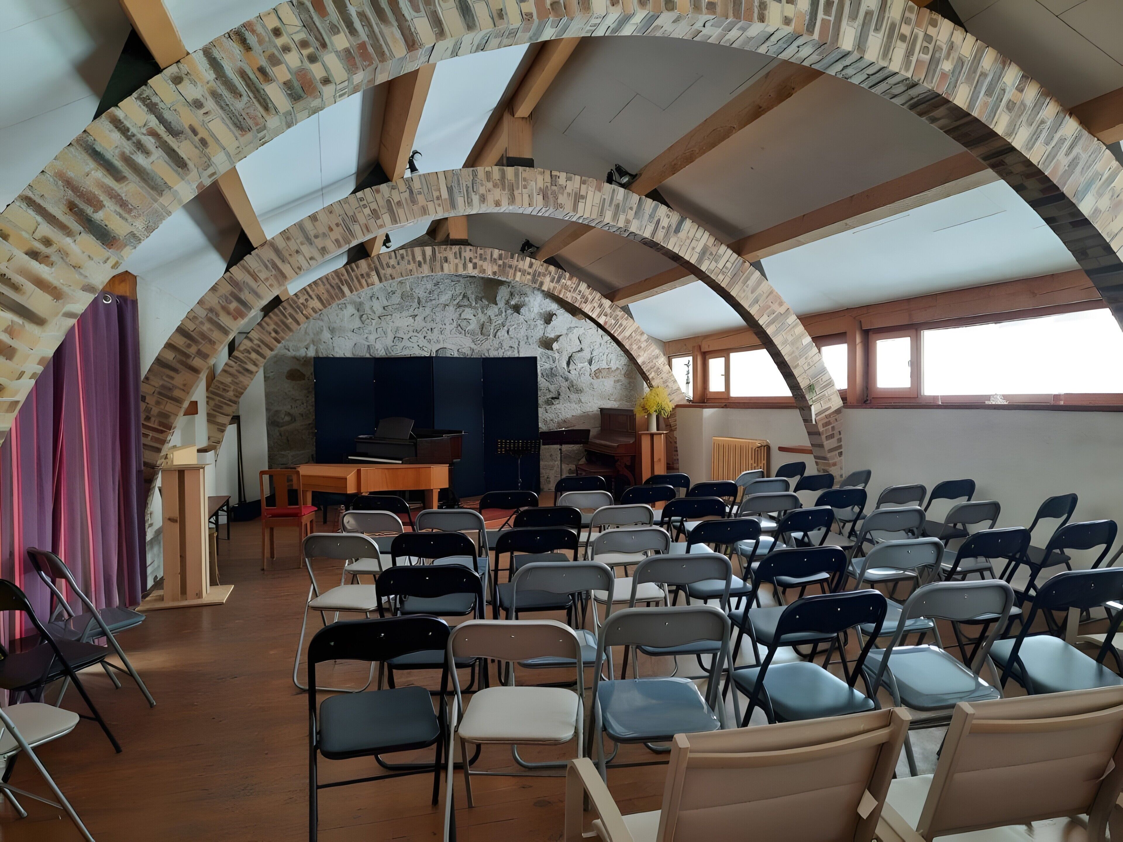 A room with a high arched ceiling, a piano, chairs arranged for an audience, and a stone wall.