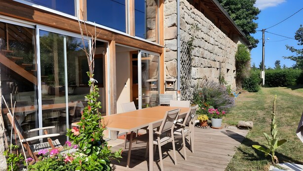 A wooden deck with a table and chairs, potted plants, and a stone building with large windows.