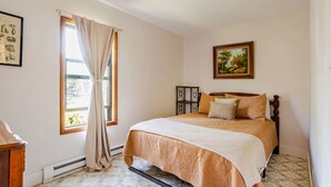 A bedroom with a bed, a window with curtains, a painting on the wall, and a radiator.