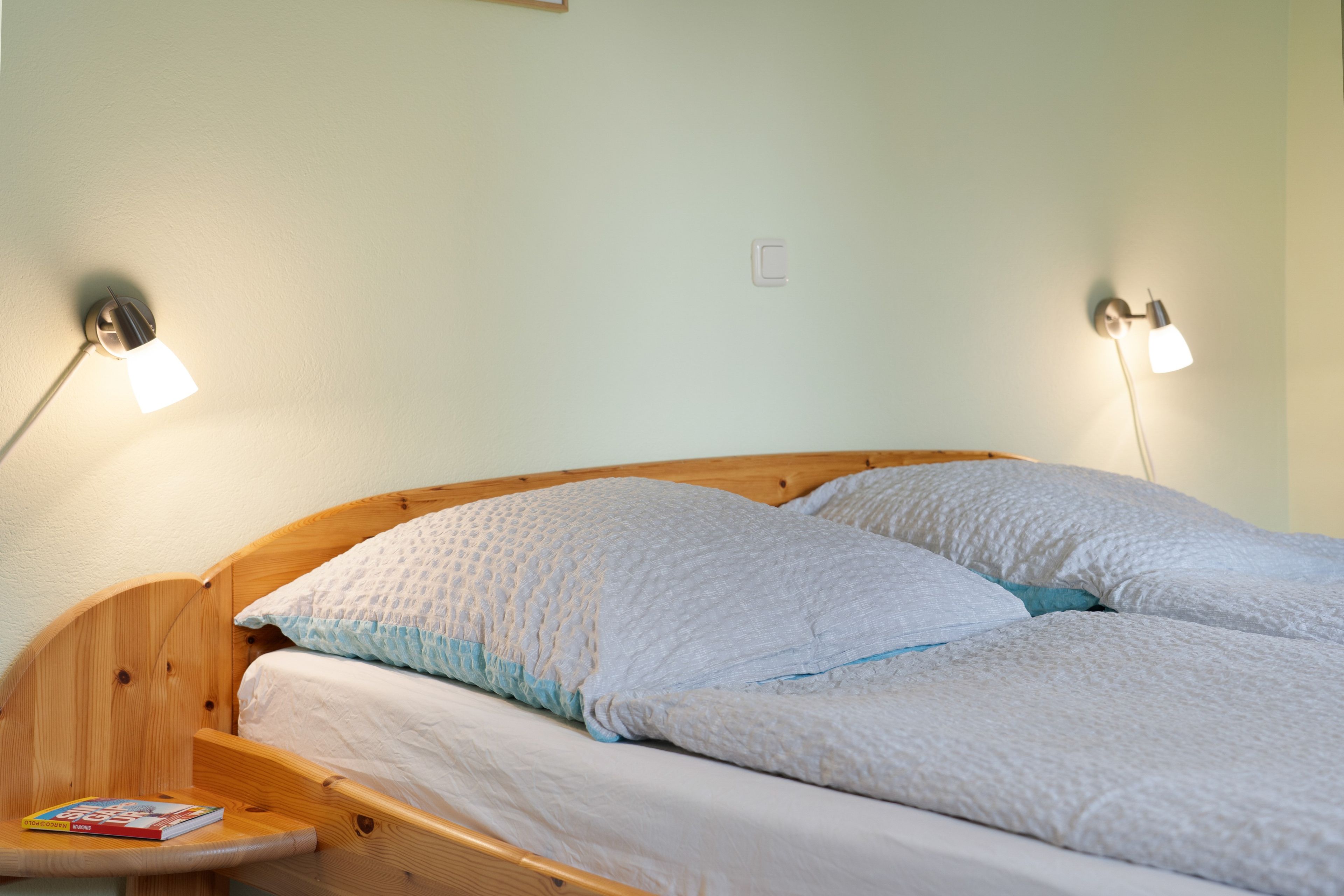 Una cama con dos almohadas, una mesita de noche con un libro y dos lámparas de pared.