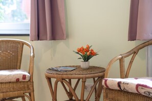 Een kamer met twee rieten stoelen, een bijzettafeltje met een bloempot en een raam met gordijnen.