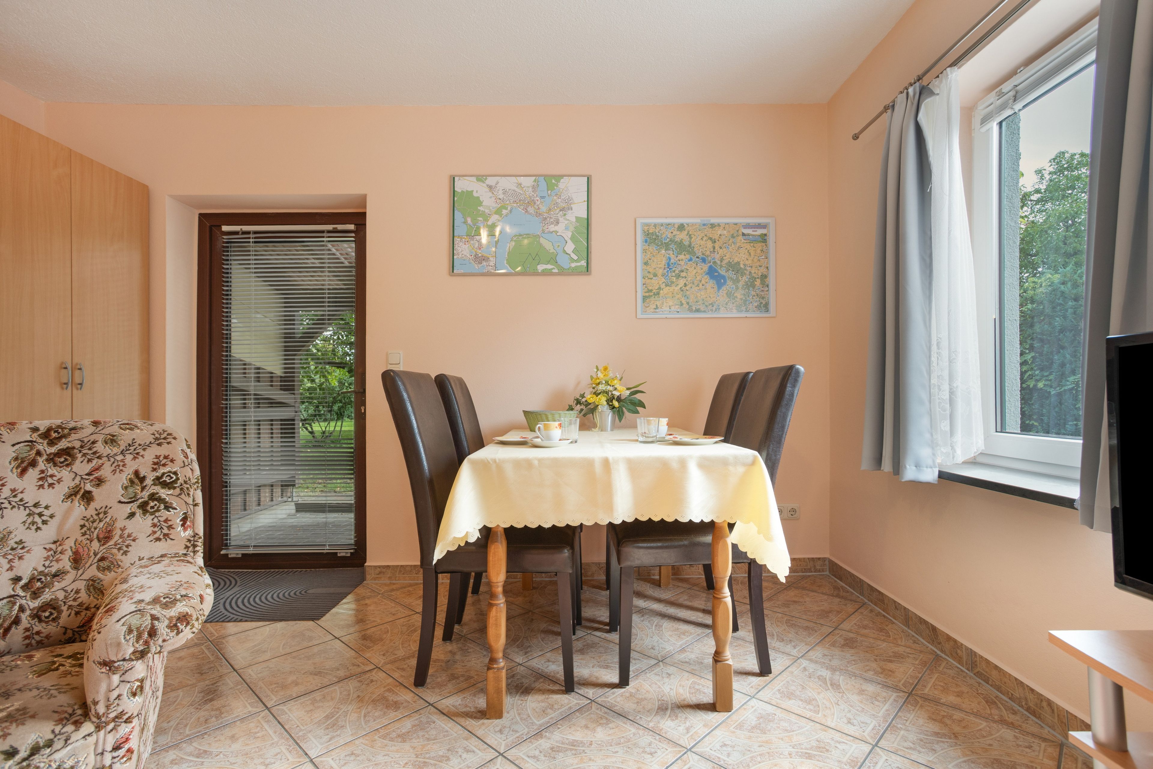 Un comedor con una mesa dispuesta para dos, dos sillas, un sofá, un ventanal con cortinas y dos mapas murales.