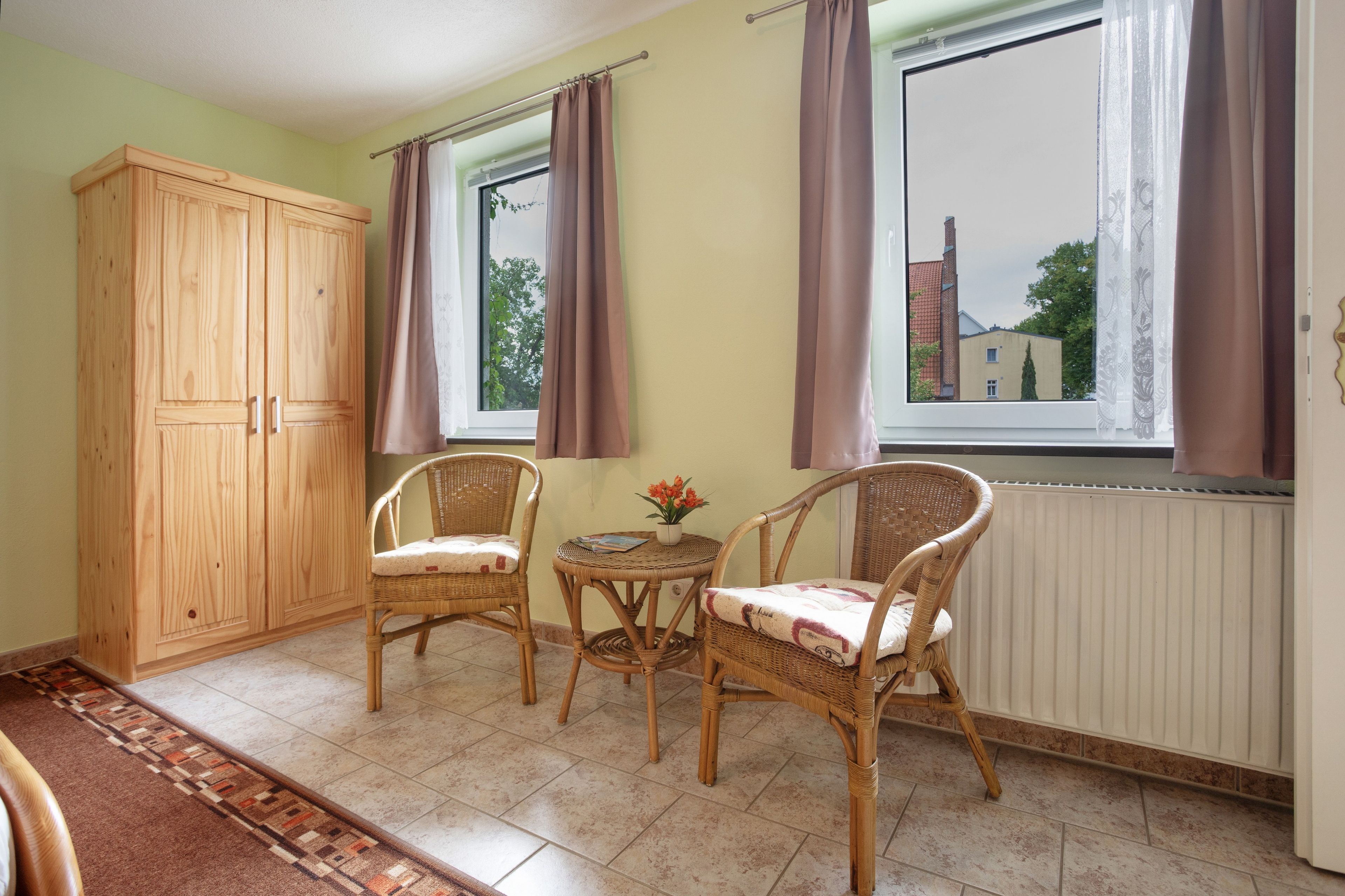 Ein Zimmer mit einem Holzschrank, zwei Korbstühlen, einem kleinen Tisch mit Blumen in einer Vase und Blick auf ein Gebäude durch ein Fenster.