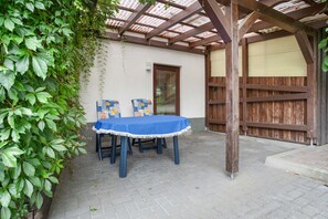 Een terras buiten met een tafel en stoelen, een houten pergola en een muur met een deur.